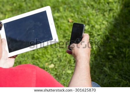 An elderly man browsing the internet with a tablet outdoor with a green grass on the background. He is also using his cell phone.