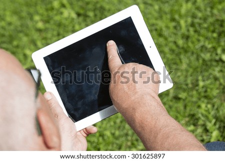 An elderly man browsing the internet with a tablet outdoor with a green grass on the background.