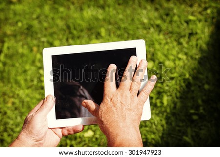 An elderly man browsing the internet with a tablet outdoor with a green grass on the background. Image has a strong vintage effect applied to create some artistic flavor.