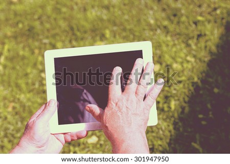 An elderly man browsing the internet with a tablet outdoor with a green grass on the background. Image has a strong vintage effect applied to create some artistic flavor.