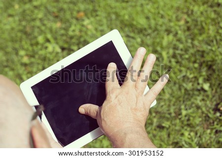 An elderly man is browsing the internet with a tablet outdoor with a green grass on the background. Image has a strong vintage effect applied.
