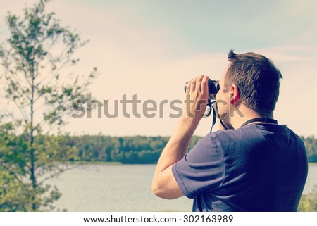 A man is watching birds and scenery with binoculars from high above the ground. The sea or lake is on the background. Image has a vintage effect applied.
