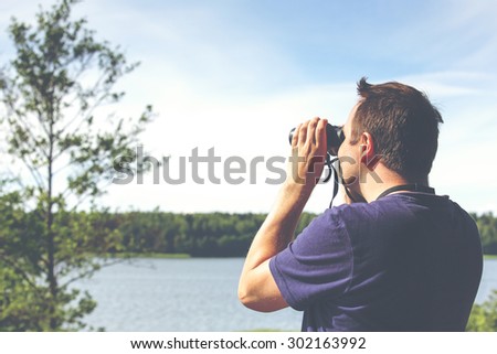 A man is watching birds and scenery with binoculars from high above the ground. The sea or lake is on the background. Image has a vintage effect applied.