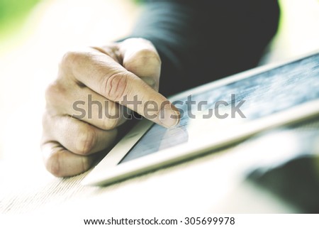 An older man is browsing the internet with a white tablet outdoor. Image has a vintage effect applied.