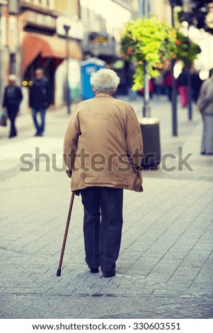 An elderly man is walking in the city with a walking stick. Some unidentified person are walking in the streets. Image has a vintage effect applied.