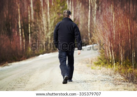 An older man is walking on the road. He is walking away. The man is wearing dark jeans and a black jacket. Image taken during spring time. Image has a vintage effect applied.