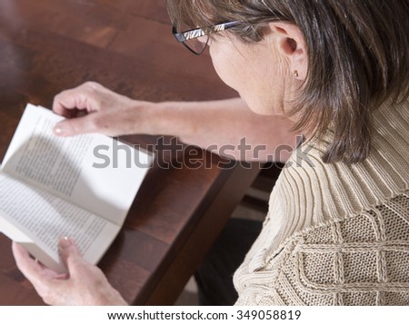 An elderly woman is reading a book on a kitchen table. Image taken against a wooden table and indoor.