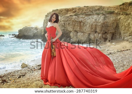 beautiful girl on the seashore at sunset in a long red dress with a violin in her hands