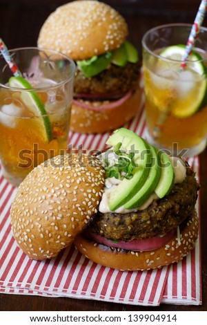 Two vegetarian sweet potato bean burgers with avocado on sesame buns, with one upper bun tilted to showcase the layers. Served with iced tea on a sheet of candy-stripe napkin