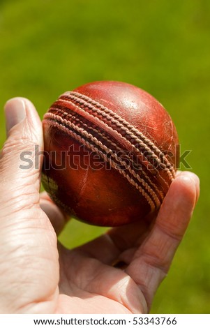 Used red leather cricket ball being held by hand against a green grass background