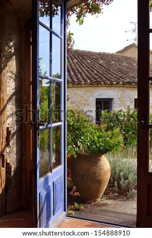 View Of An Old Stone Building Through A Blue French Door, With A Charming Urn.
