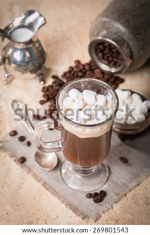 Glass Cup of coffee with marshmallow, milk jug, spoon and coffee beans in jar on stone background
