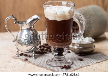 Glass Cup of coffee with marshmallow, milk jug, spoon and coffee beans in jar on stone background