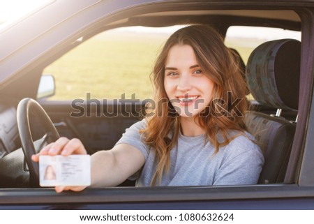Smiling young female with pleasant appearance shows proudly her drivers license, sits in new car, being young inexperienced driver, looks with joyful expression. I get it finally! Successful woman