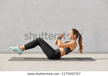 Self determined sporty woman with pony tail, dressed in leggings, top, sneakers, smartwatch makes on press, wants to have muscular body, has workout in in gym. People, gym and sport concept.