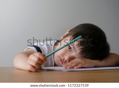 Portrait Kid boy holding colour pencil sitting alone and looking out with bored face,Preschool child laying head down on table with sad face,Five years old kid bored with school homework,spoiled child