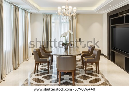 Front view of stylish and light dining room with big windows and crystal chandelier in center of ceiling. Luxury interior of big room with wooden table in center and armchairs around.
