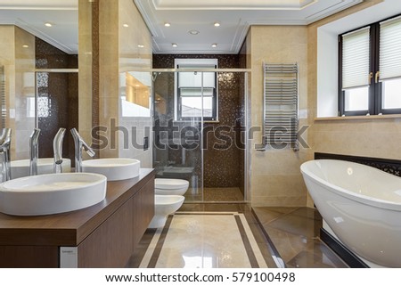 Big and stylish bathroom in brown and beige colors, with ceramic bath under window, big mirror on wall, two sink and bowl. Shower cabin with glasses wall.