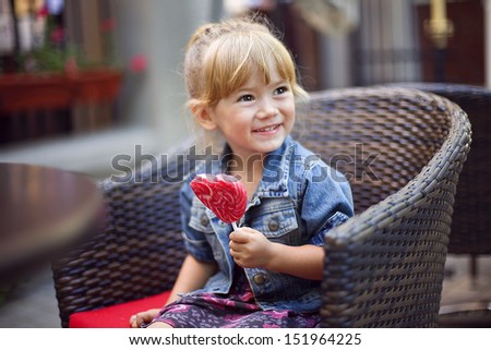 cute little girl holding a big heart shaped lollipop in the cafe outdoors