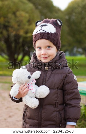 Toddler boy in brown coat and knitted bear hat with toy bear in his hands outdoor in autumn