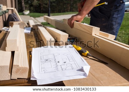 Carpenter working with technical drawing or blueprint construction paper lying on a workshop outdoor desk, surrounded with carpentry tools and wood, furniture making or house renovation process
