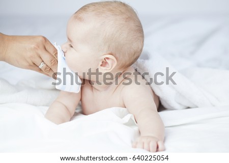 Drooling baby or running nose getting wiped by mom\'s hand, lying naked in white sheets