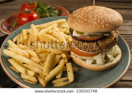 Healthy burger alongside french fries on a decorative plate with tomatoes and fried eggplant in the background