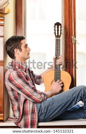 Looking for inspiration. Handsome young men sitting on the windowsill and holding an acoustic guitar in his hand