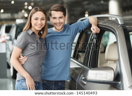 We Have Made The Right Choise. Handsome Young Men Standing Near The Car At The Dealership Hugging His Girlfriend And Holding A Key