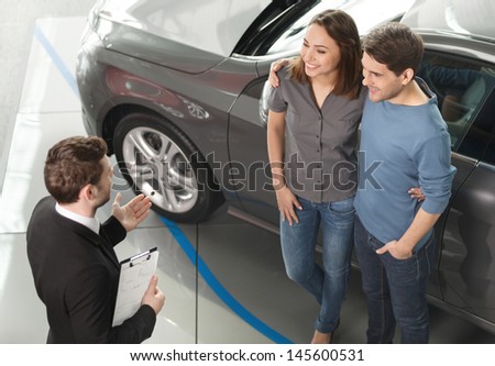 Buying their first car together. High angle view of young car salesman standing at the dealership telling about the features of the car to the customers