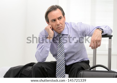 Tired of traveling. Portrait of a thoughtful senior businessman sitting in a sofa at the hotel room and holding his hand on the suitcase