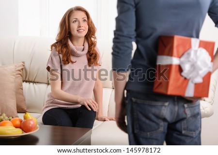 Surprise for her. Beautiful young woman sitting at the couch while her boyfriend holding a gift behind his back