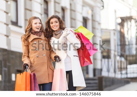 She knows where are the best prices. Two beautiful young women standing with shopping bags in their hands while one of them pointing away
