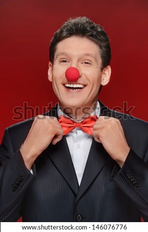Funny man. Portrait of cheerful man with a clown nose touching his bow tie and looking at camera