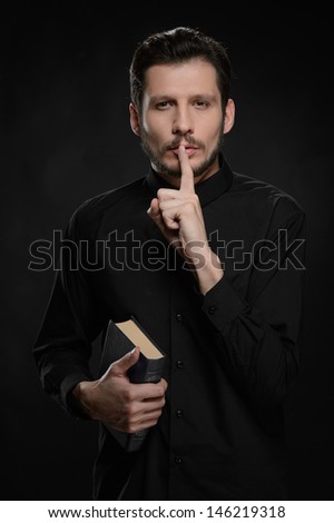 Asking to keep silence. Portrait of priest holding his hand against lips while standing against black background