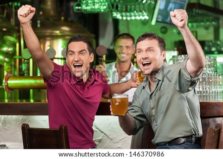 Soccer Fans At The Bar. Two Happy Football Fans Cheering At Bar And Drinking Beer While Bartender Serving Beer At The Background