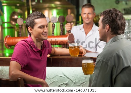 Talking at bar. Two cheerful male friends talking at bar and drinking beer while bartender serving beer at the background