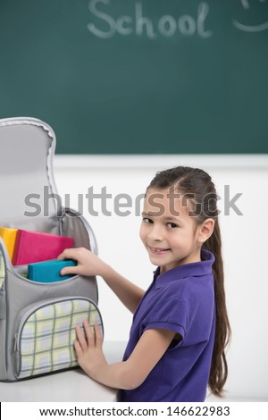 Packing her book bag. Cheerful little schoolgirl packing her book bag and smiling at camera