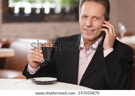 Talking to business partner. Cheerful businessman drinking coffee and talking at phone while sitting at restaurant