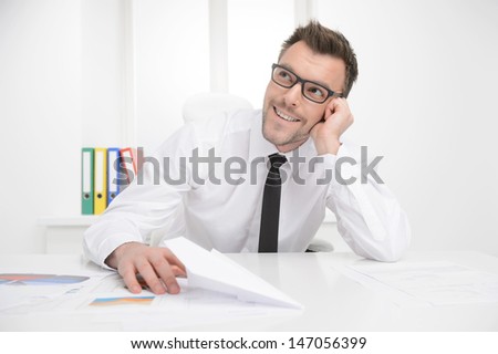 Dreaming of vacation. Portrait of thoughtful businessman sitting at his working place and holding a paper plane in his hand