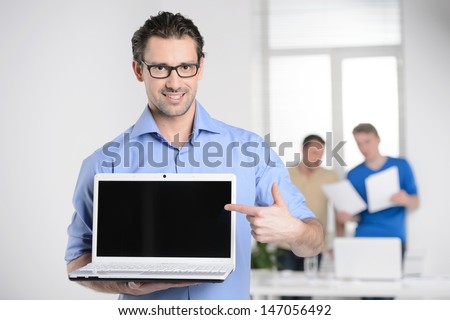 Men With Laptop. Happy Young Businessman Holding A Laptop And Pointing It While Colleagues Working On The Background