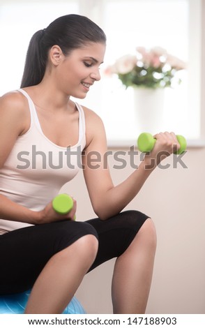 Women weightlifting. Beautiful young women exercising at home while sitting on the fitness ball and using the dumbbells