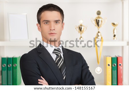 Confident businessman. Confident young businessman standing in front of his business trophies and holding his arms crossed