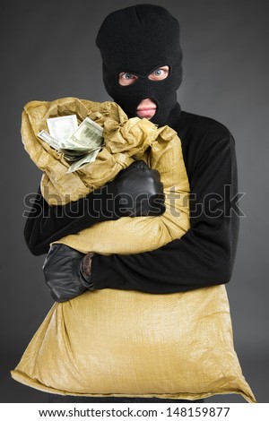 Thief with stolen goods. Front view of frustrated men in black balaclava holding a stolen money bag while standing isolated on grey