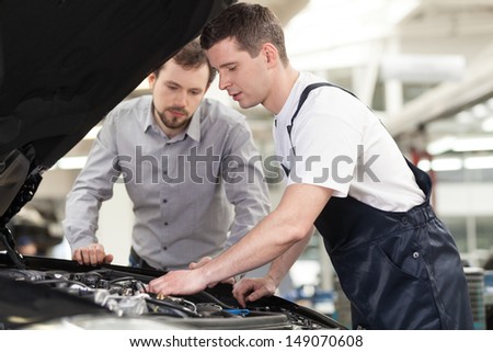 Examining a car Confident auto mechanic examining car engine while customer looking at it Examining a car Confident auto mechanic examining car engine while customer looking at it