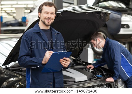 Mechanics At Repair Shop. Cheerful Young Mechanic Writing Something In His Clipboard While Another One Fixing A Car On The Background