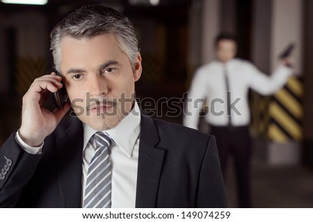 Killer and victim. Confident mature businessman talking at mobile phone while another men with gun standing behind him