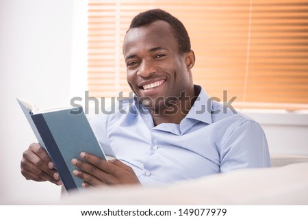 Reading an exciting book. Cheerful African descent reading a book and smiling at camera