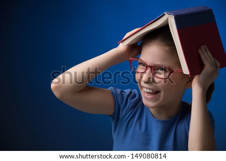 Happy schoolgirl. Happy little girl in glasses carrying a book on head while standing isolated on coloured background