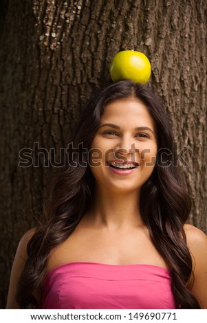 Apple on her head. Cheerful young woman standing back to the tree and holding apple on her head